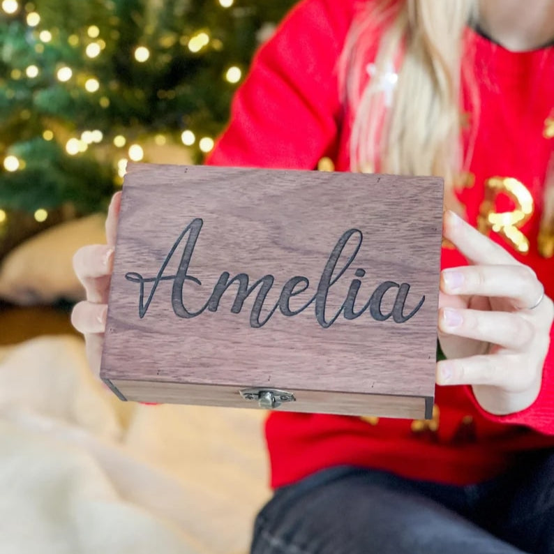 Person holding a wooden sign with 'Amelia' engraved, Christmas tree in the background