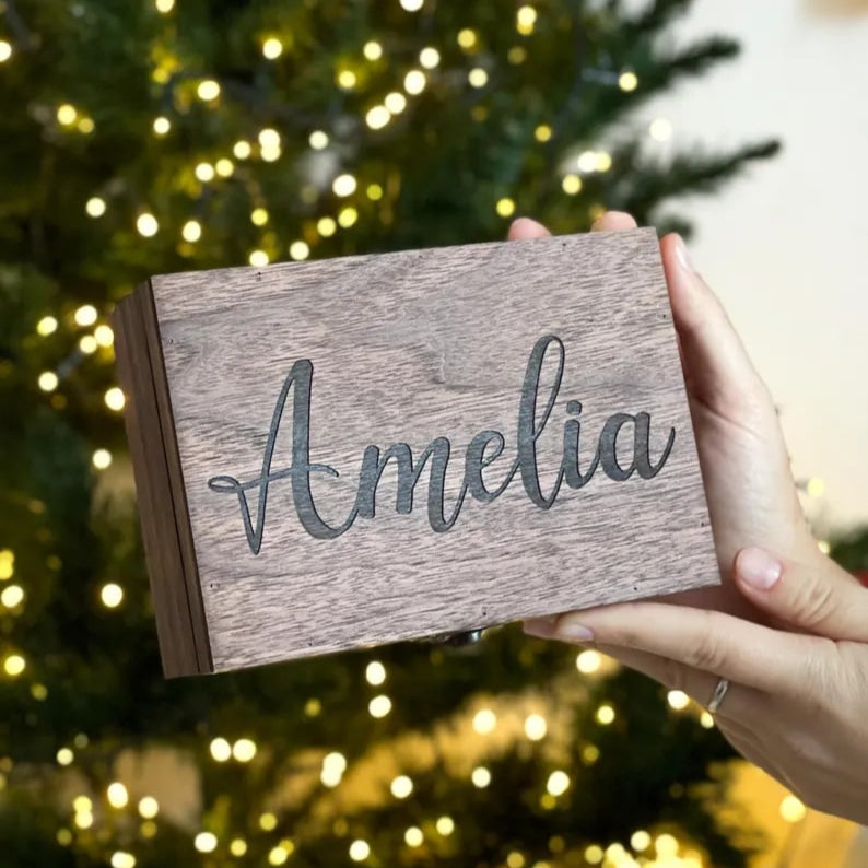 Hand holding a wooden box with 'Amelia' engraved, Christmas tree in the background
