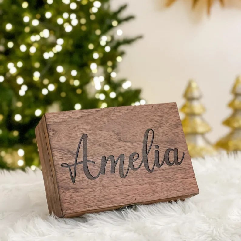 Wooden block with 'Amelia' engraved, placed on a fluffy white surface with a blurred Christmas tree in the background.
