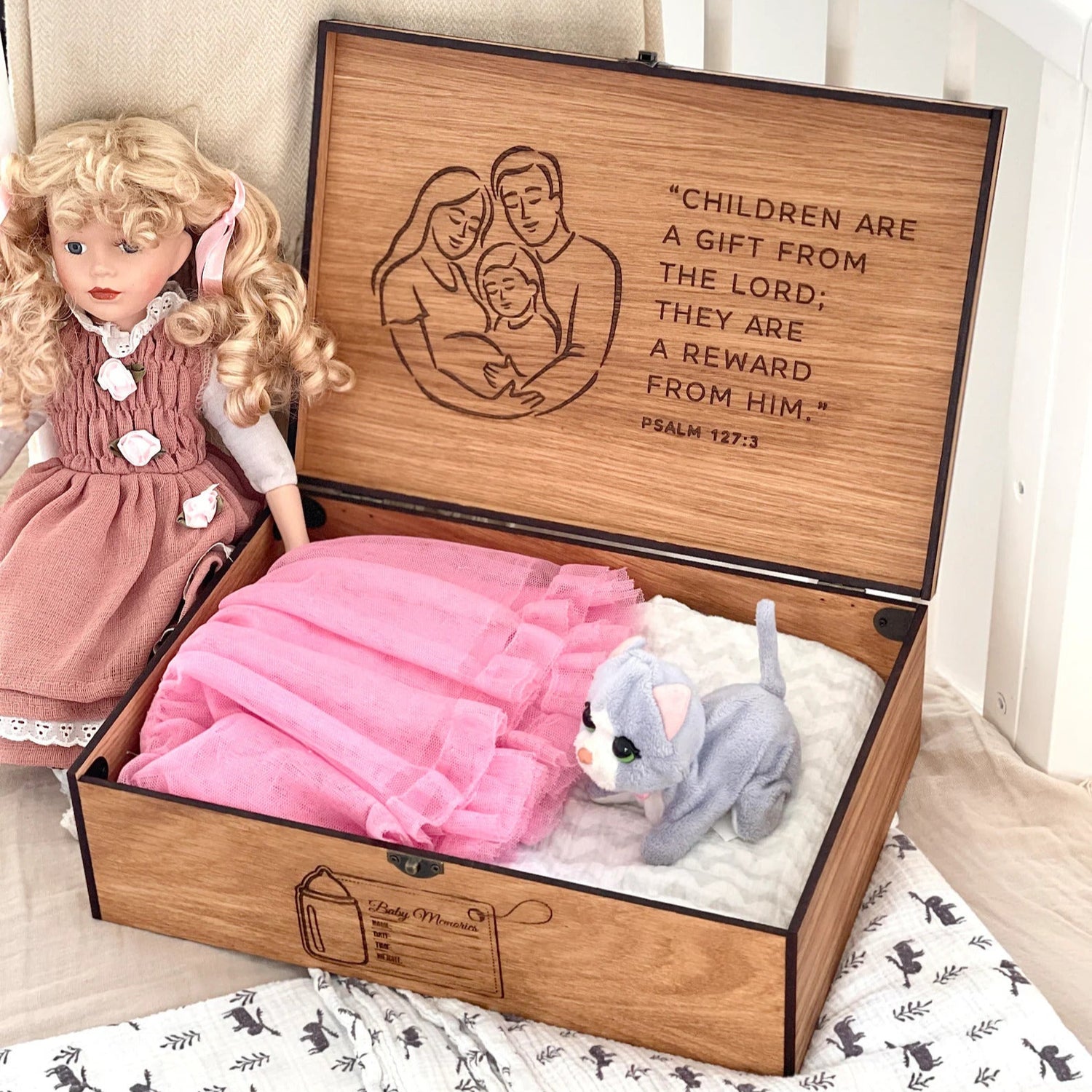 Wooden toy box with engraved text, doll, and teddy bear on a patterned rug.