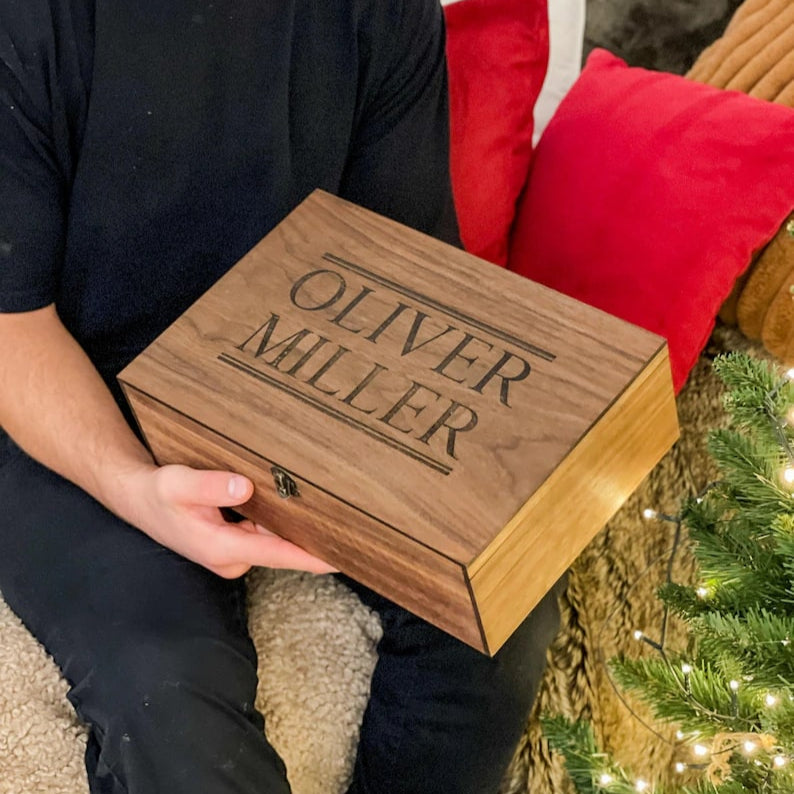 Person holding a wooden box with 'Oliver Miller' engraved, sitting on a couch with Christmas decorations.