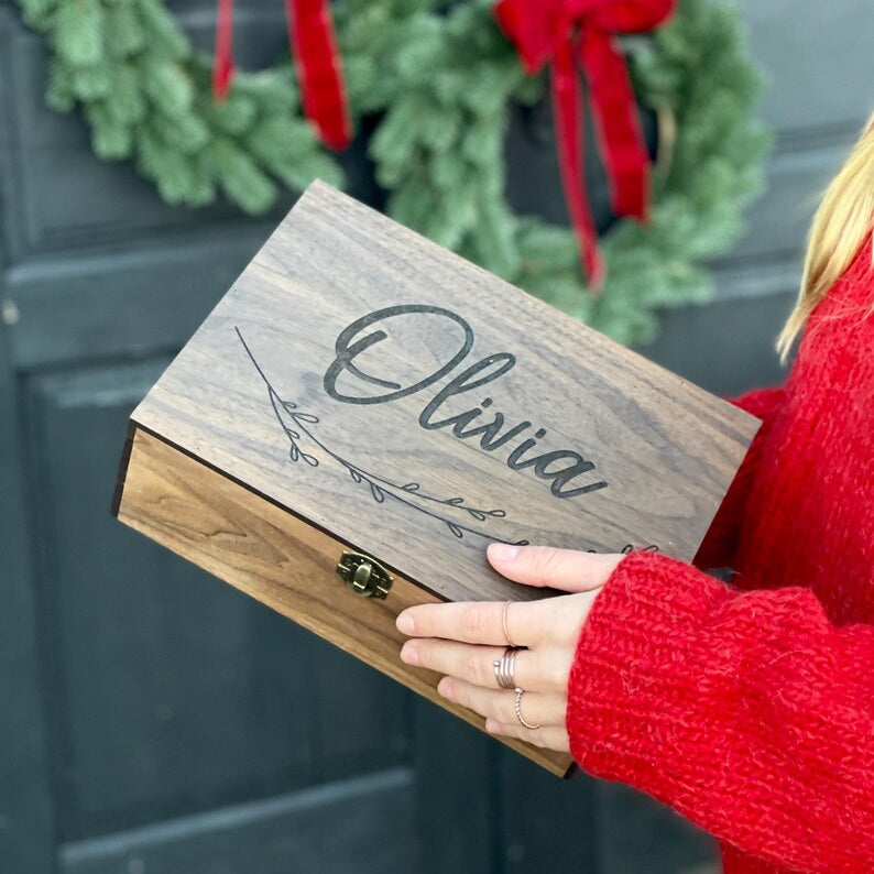 Person holding a wooden box with 'Olivia' engraved, in front of two Christmas wreaths on a door.