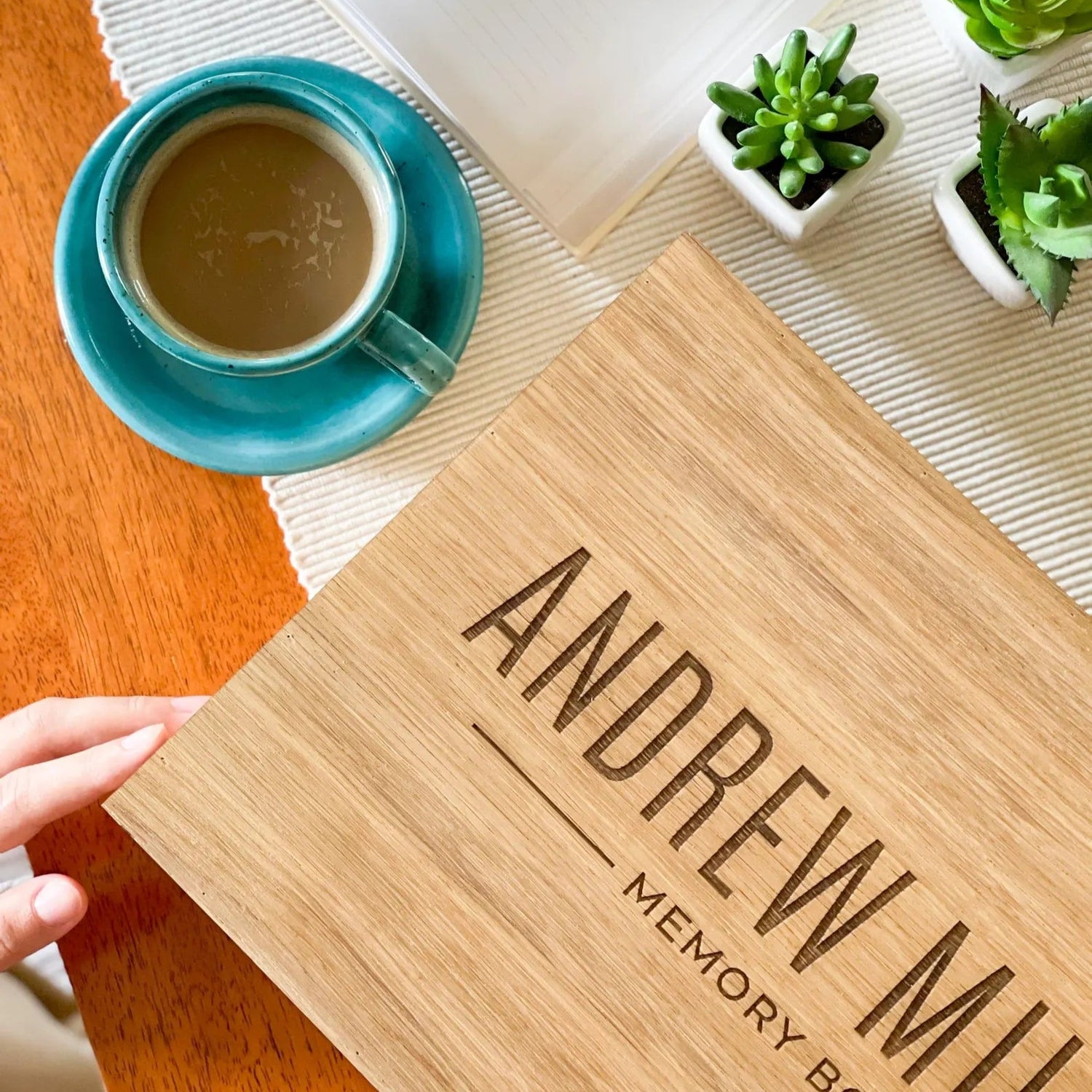 Wooden memory box with 'Andrew Miller' engraving on a table with a cup of coffee and plants.