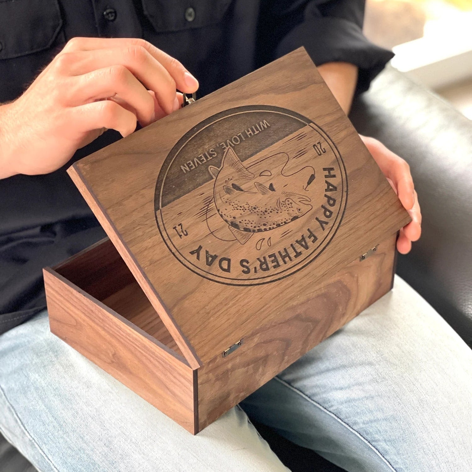 Person holding a wooden box with engraved text and design, sitting on a couch.