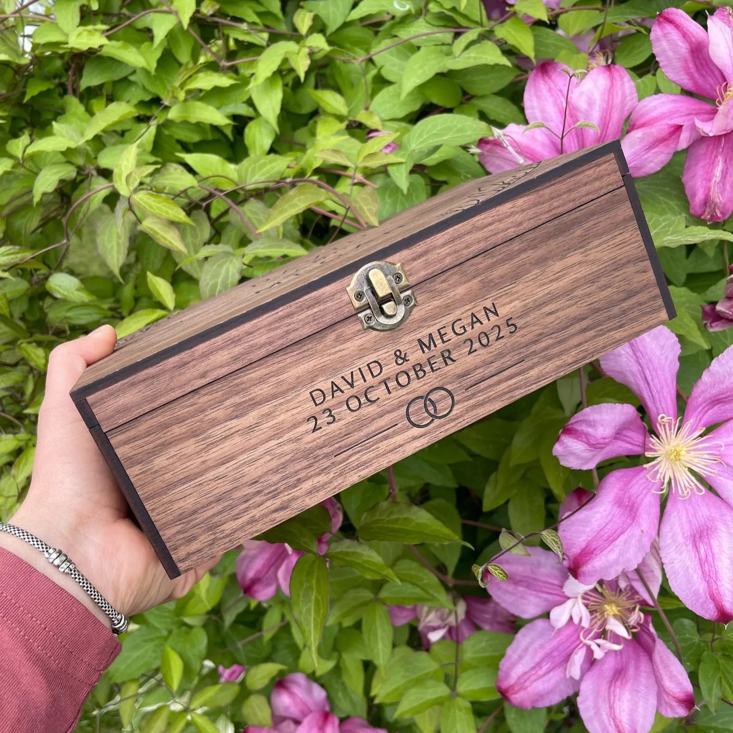 Wooden box engraved with 'DAVID & MEGAN 23 OCTOBER 2023' held in front of pink flowers.