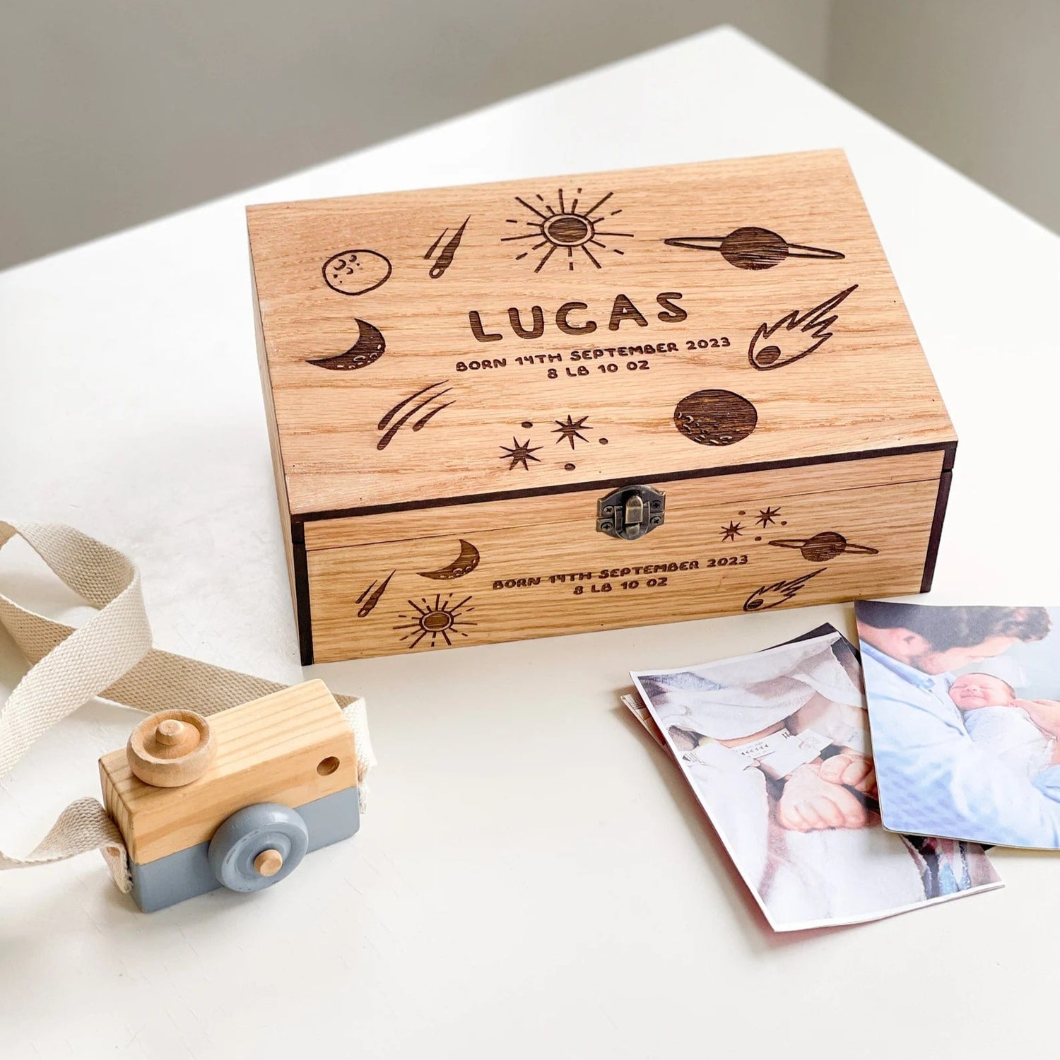 Wooden box engraved with 'Lucas' and celestial designs, accompanied by a toy camera and photos on a light surface.