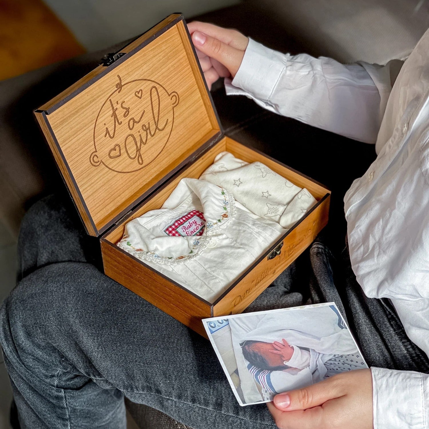 Person holding a wooden box with baby clothes and a card, sitting on a couch.