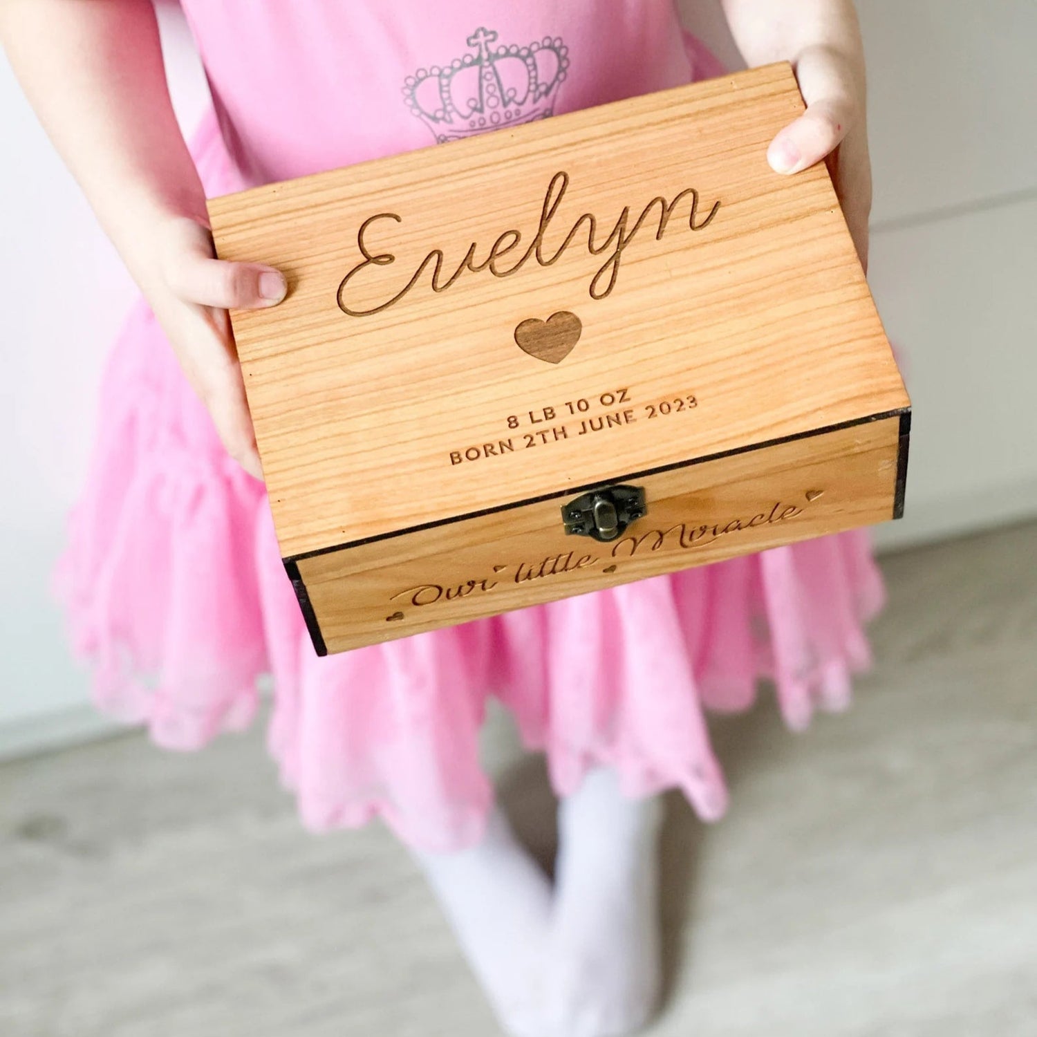 Child in a pink dress holding a wooden box engraved with 'Evelyn' and birth details.