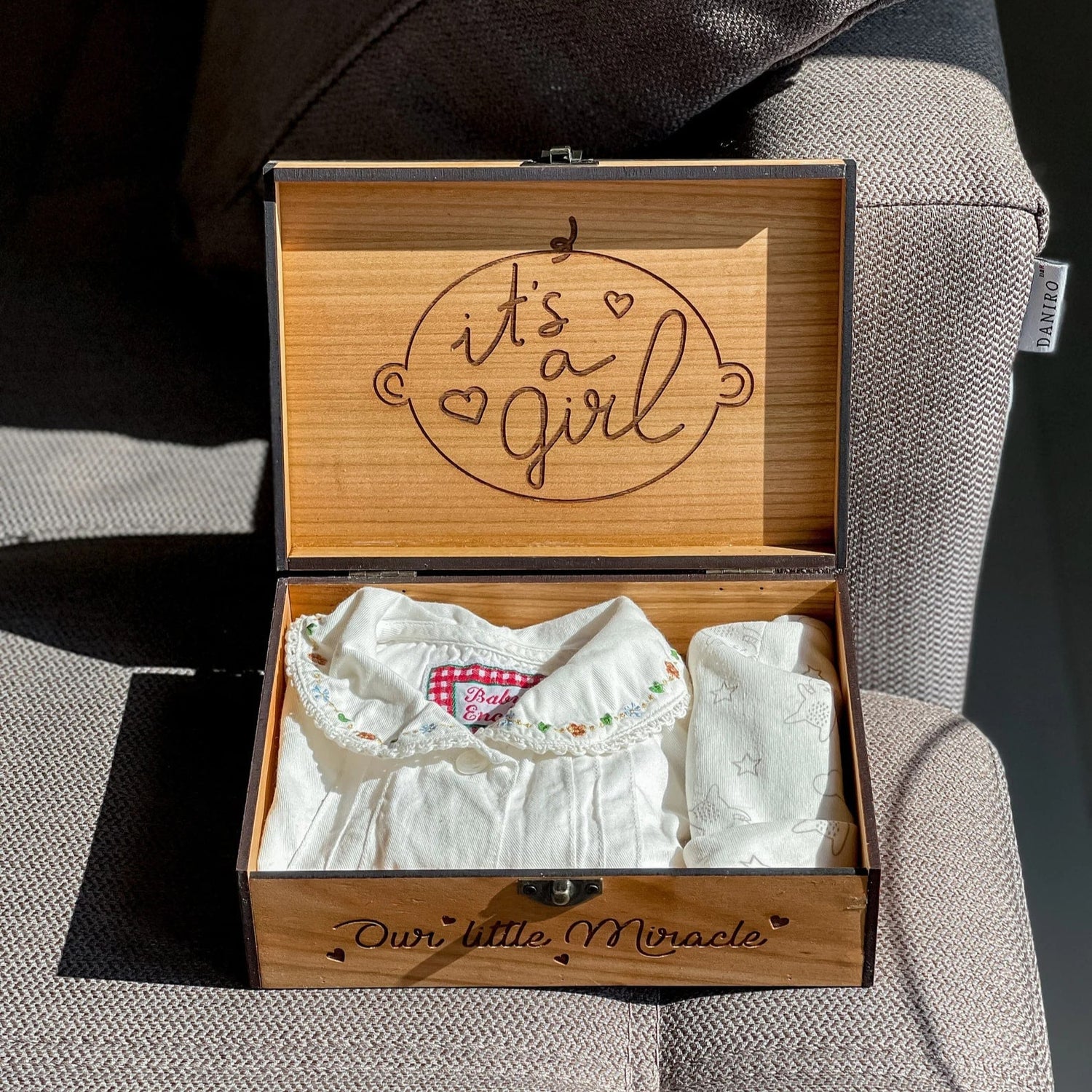 Wooden box with 'it's a girl' engraving containing baby clothes on a textured surface.