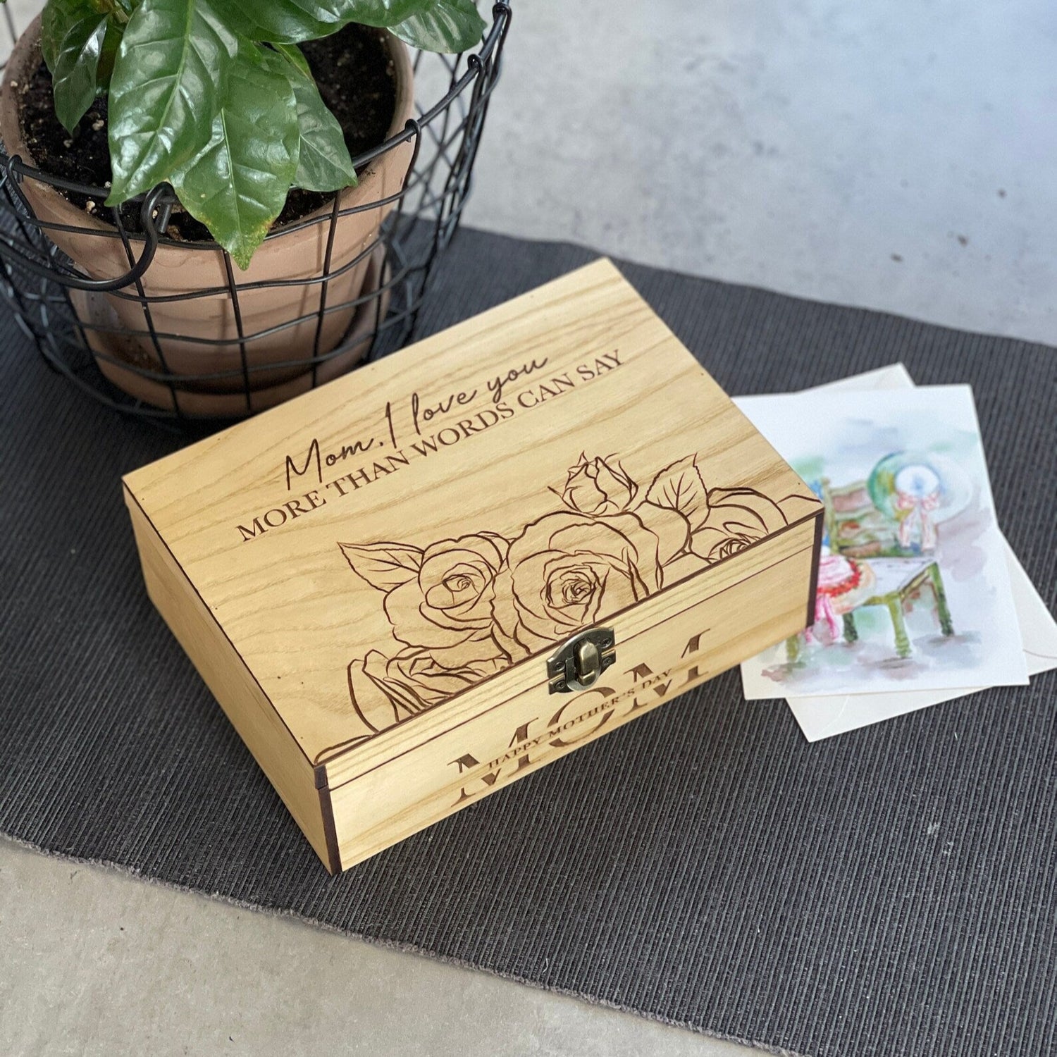 Wooden box with engraved message and floral design on a table with a plant and card in the background.