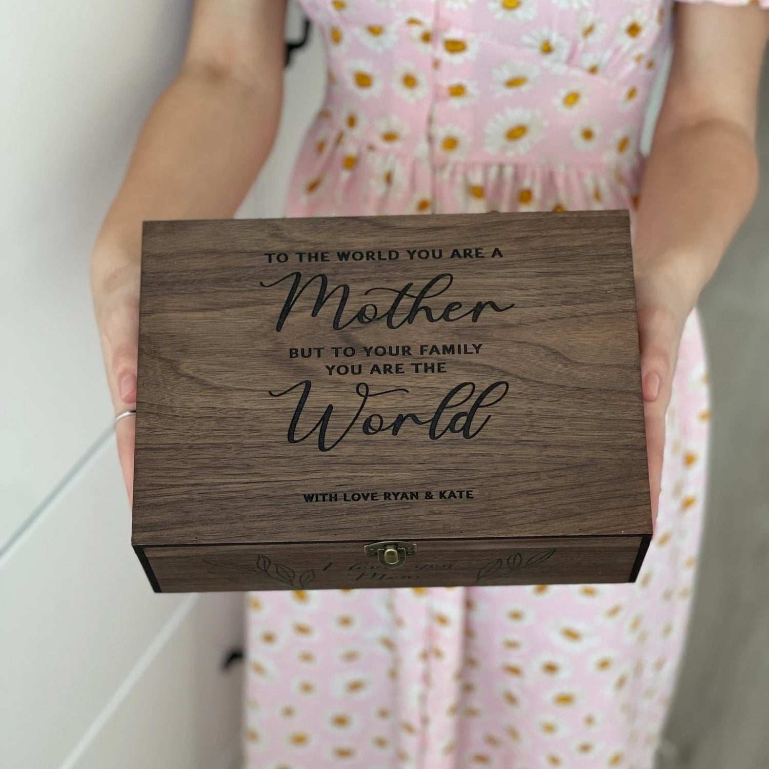 Person holding a wooden box with engraved text, wearing a pink dress with yellow flowers.