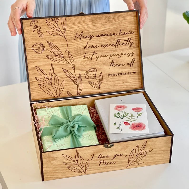 Wooden gift box with floral designs and text, containing a green bow and a card, on a white surface.