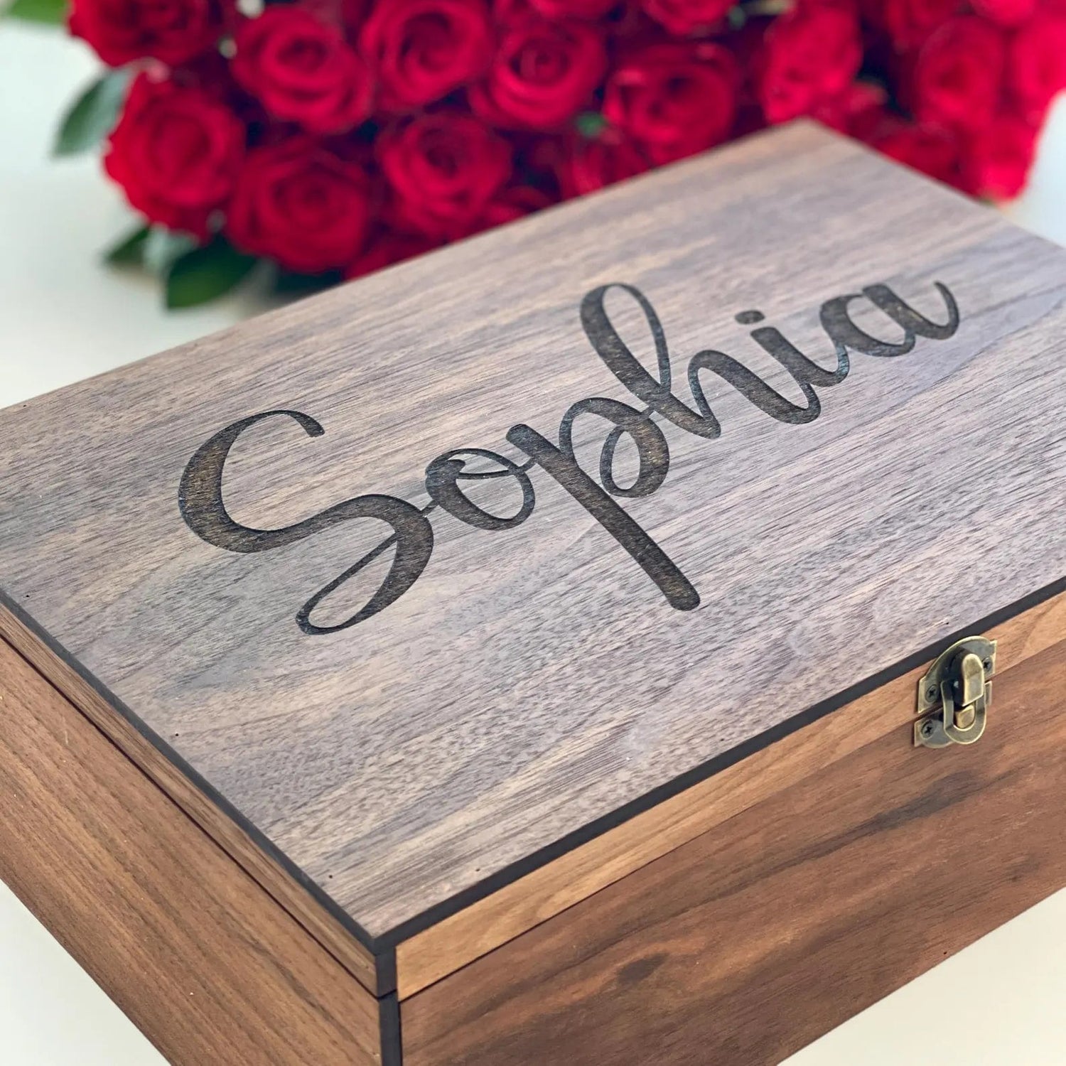 Wooden box with 'Sophia' engraved on it, in front of a bouquet of red roses.