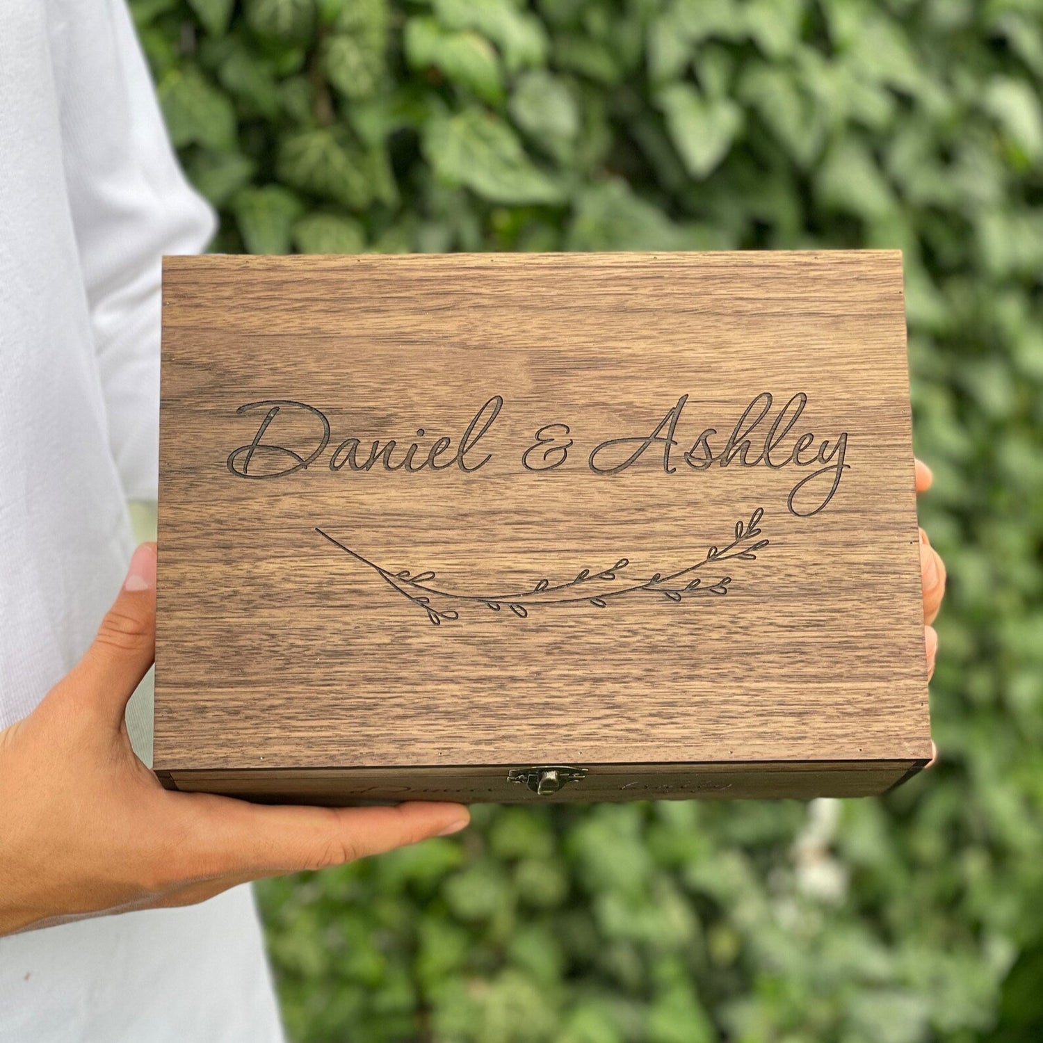 Wooden box with 'Daniel & Ashley' engraving held by a person against a green leafy background