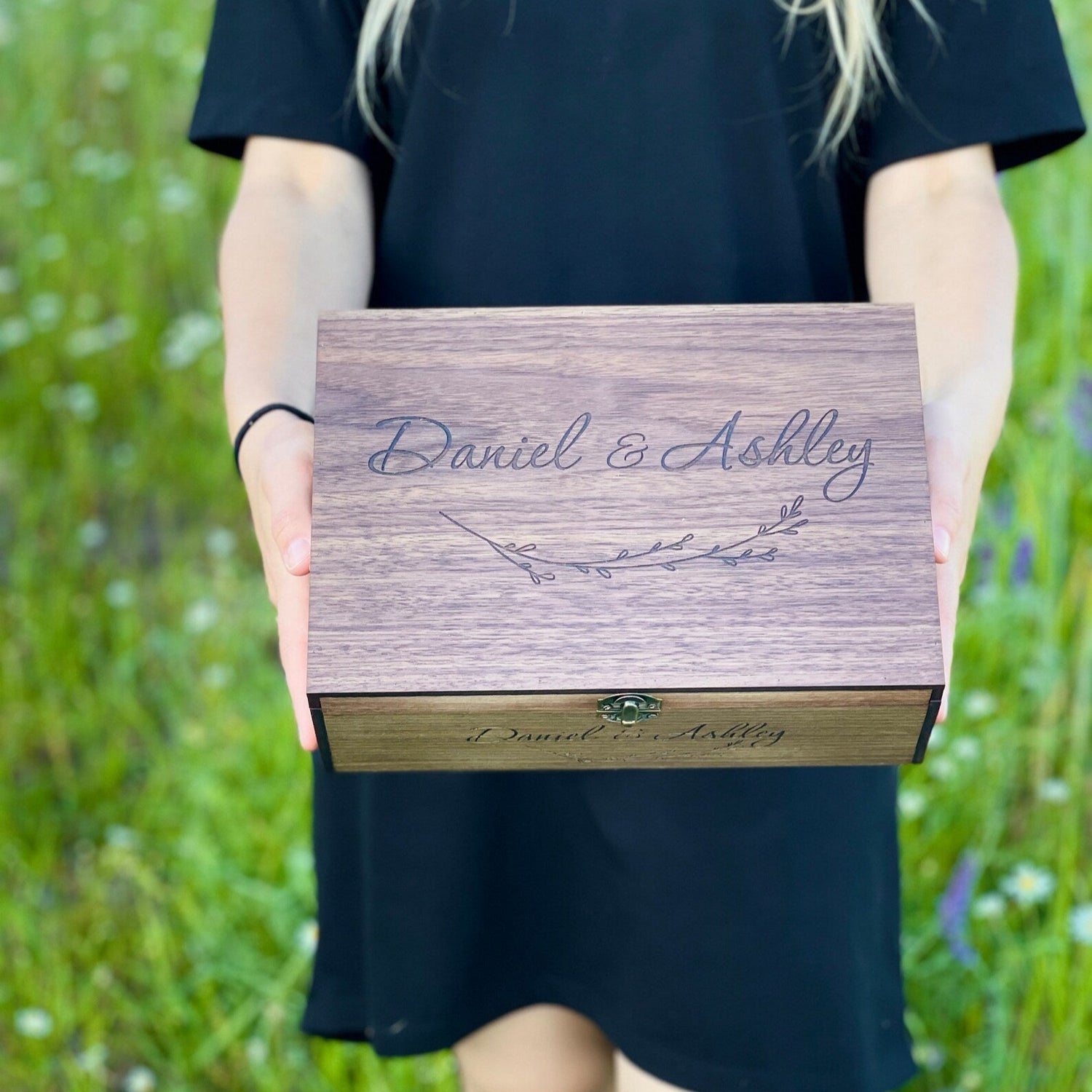 Person holding a wooden box with 'Daniel & Ashley' engraved on it, standing in a grassy field.
