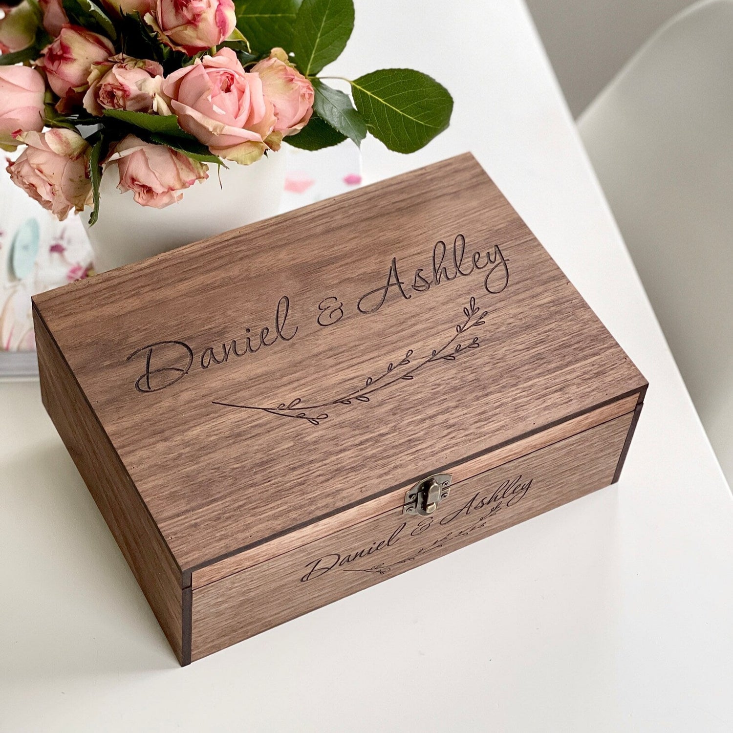 Wooden box with engraved names 'Daniel & Ashley' on a white surface with pink flowers.