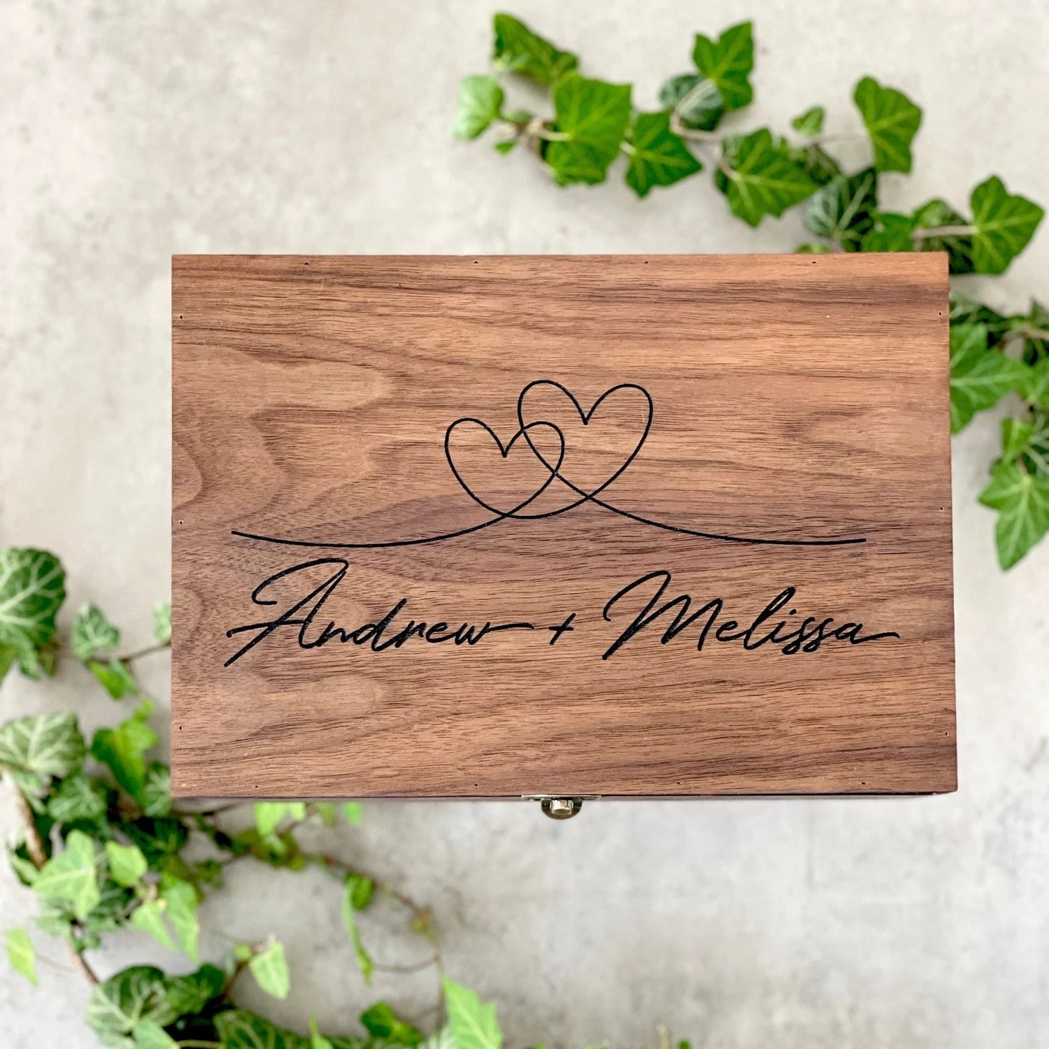 Wooden box with 'Andrew + Melissa' engraving on a white background with greenery