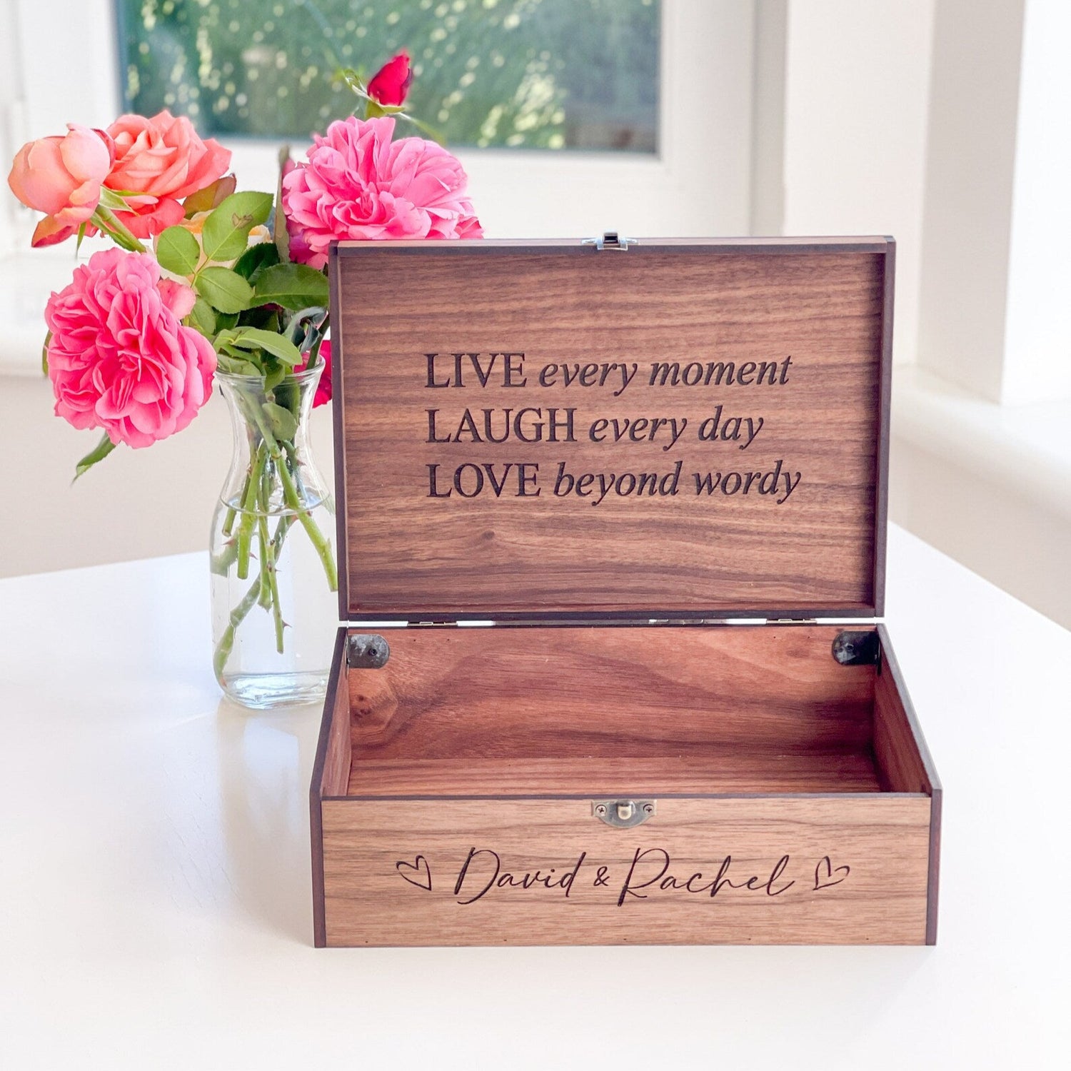 Wooden box with engraved text and a vase of pink flowers on a white surface.