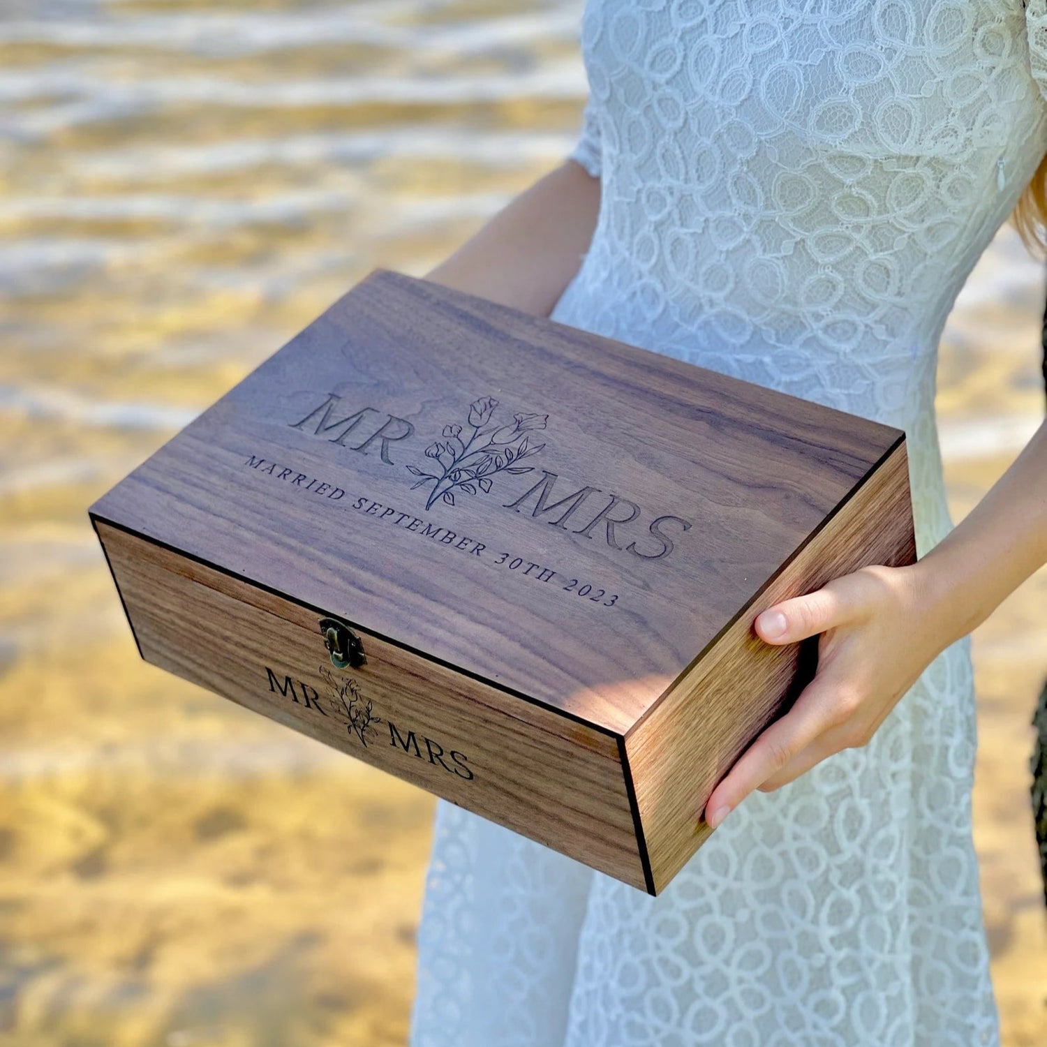 Wooden box engraved with 'MR & MRS' held by a person in a white dress by a body of water.