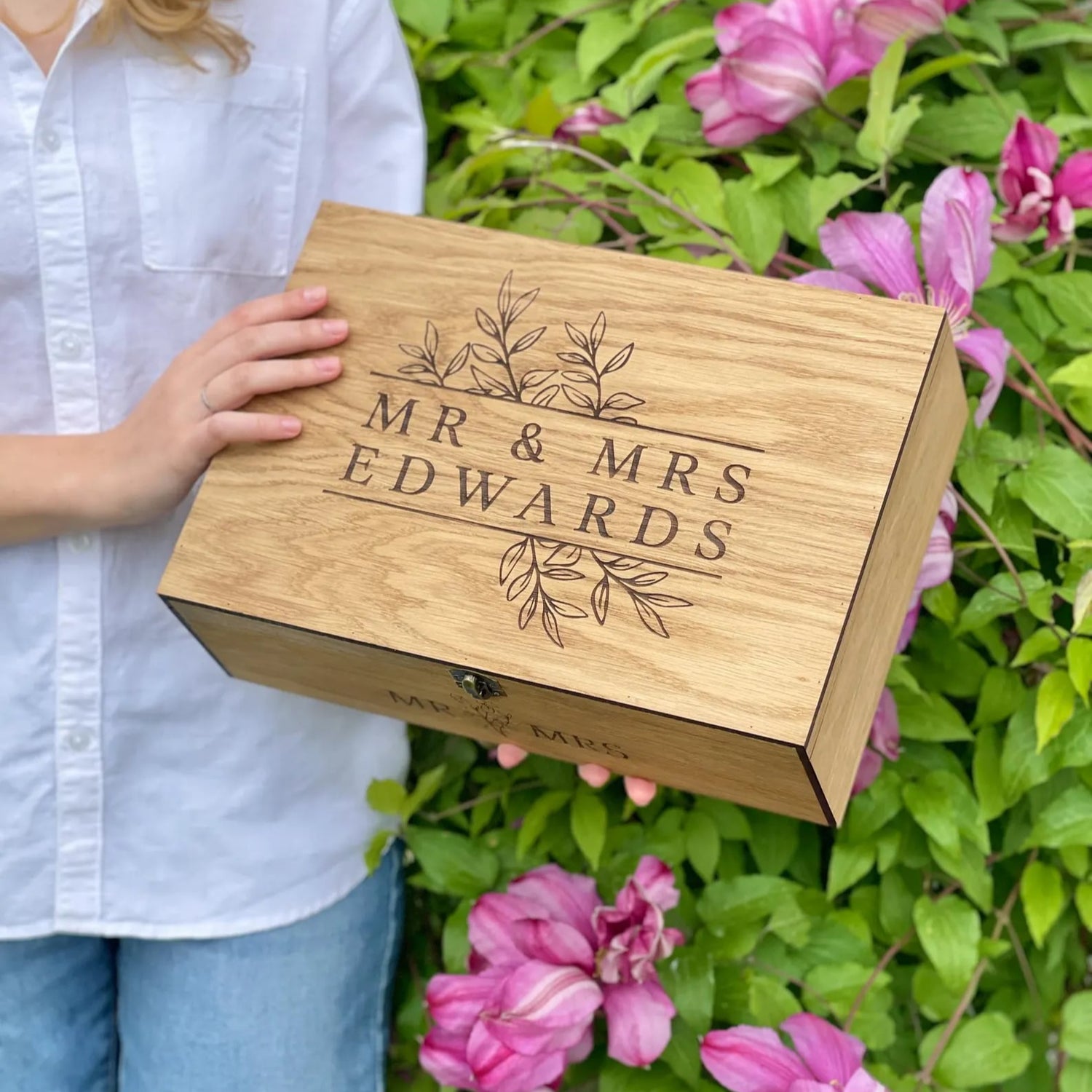 Person holding a wooden box engraved with 'Mr & Mrs Edwards' amidst pink flowers and greenery.