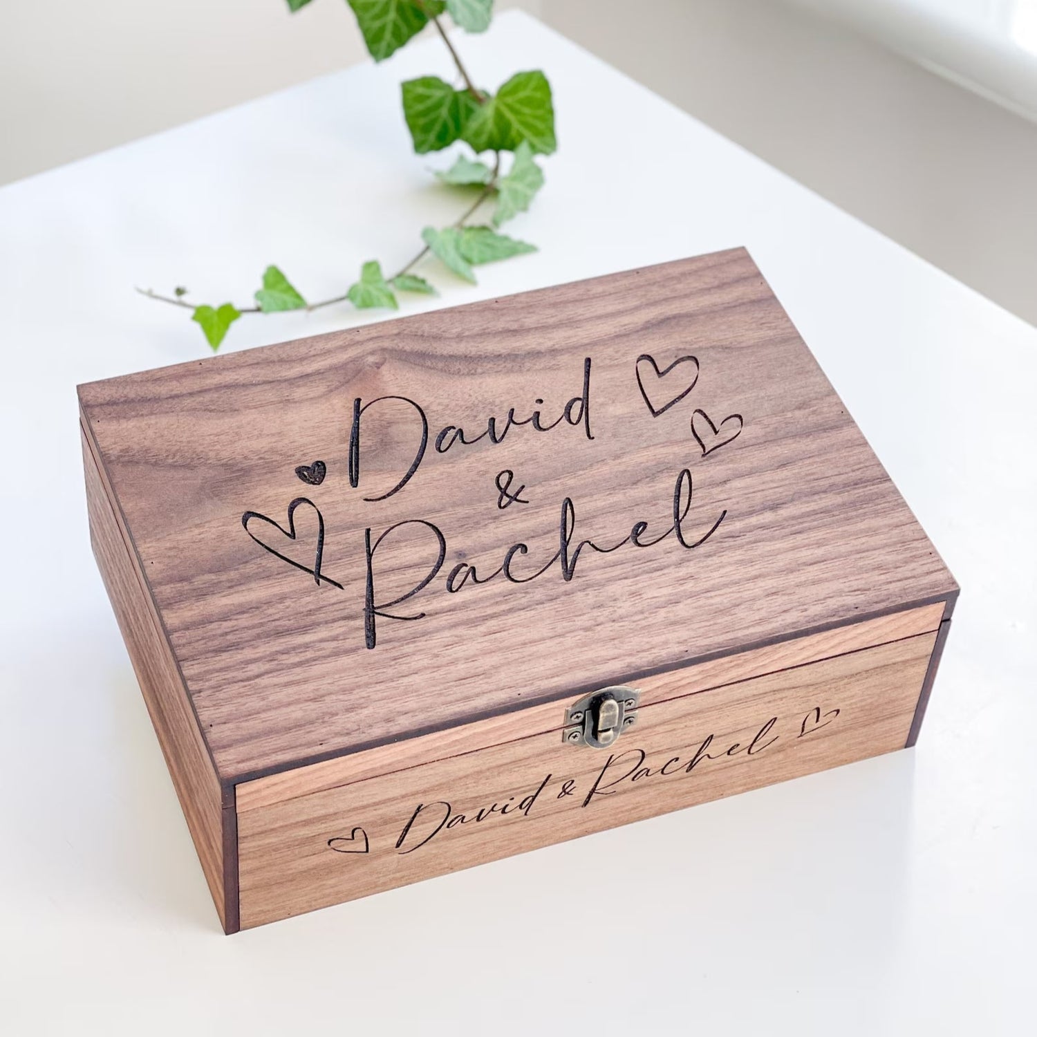 Wooden box with engraved names 'David & Rachel' on a white surface with greenery.