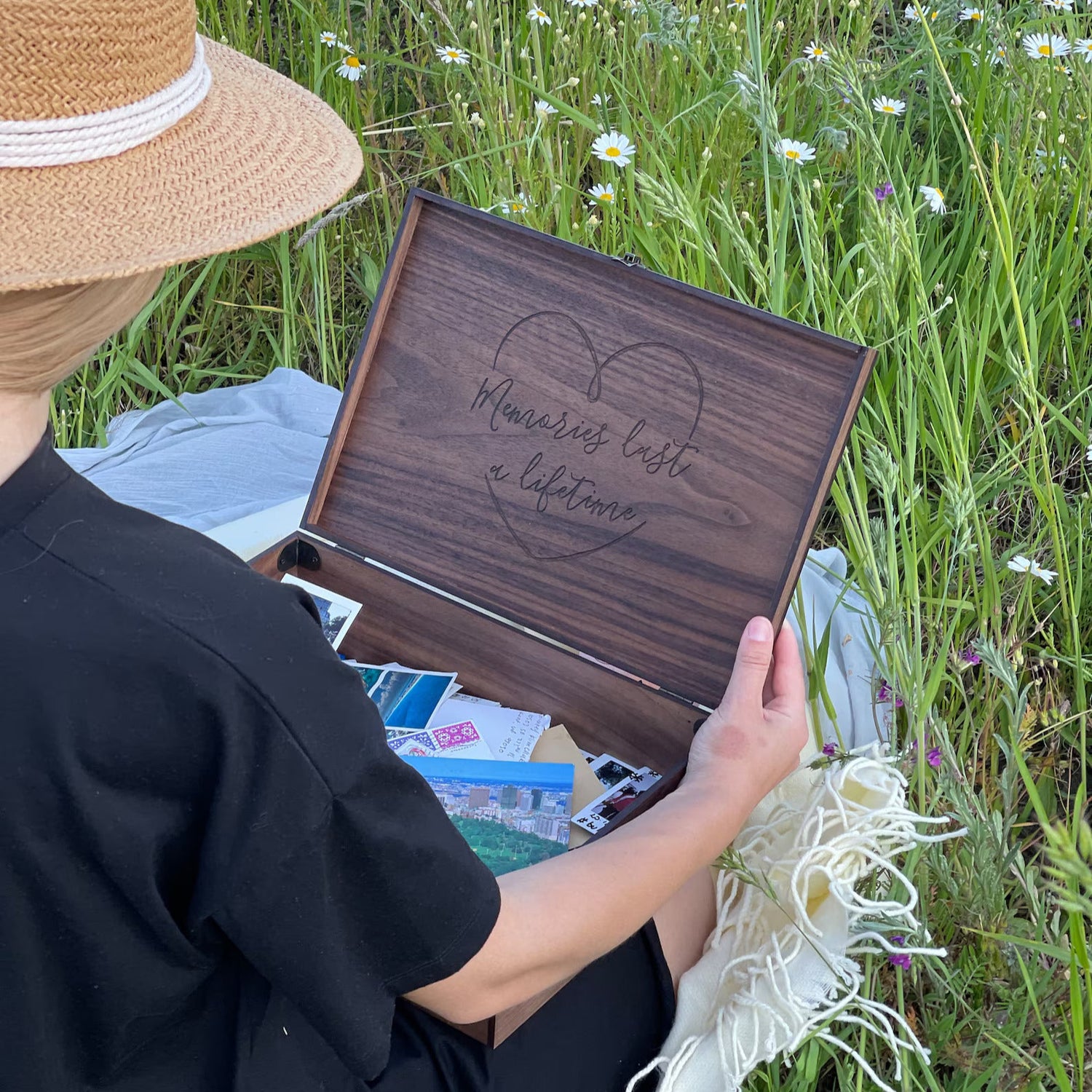Person holding a wooden box with 'Memories Last a Lifetime' engraving in a grassy field with flowers.