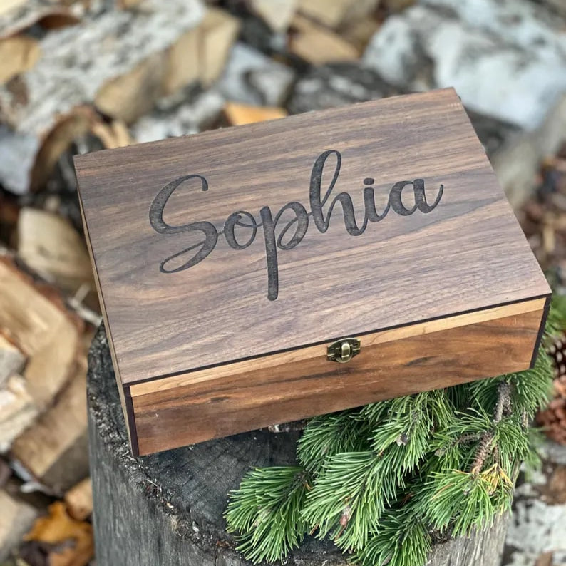 Wooden box with 'Sophia' engraved on it, placed outdoors with logs and greenery in the background.