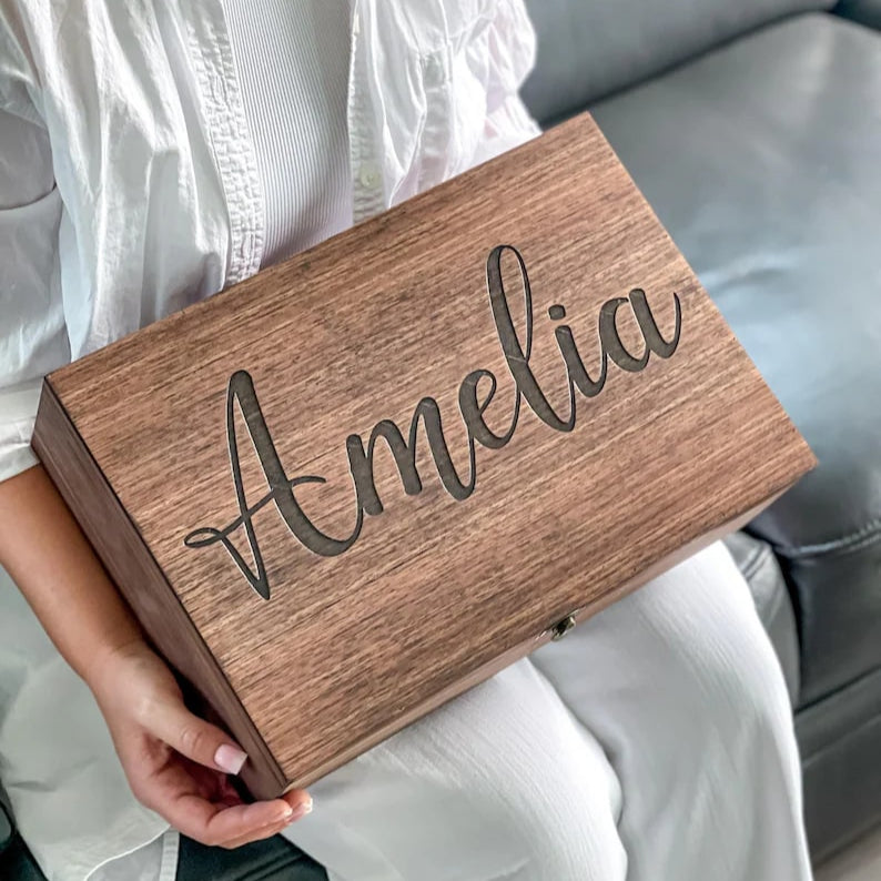 Person holding a wooden box with 'Amelia' engraved on it, sitting on a couch.