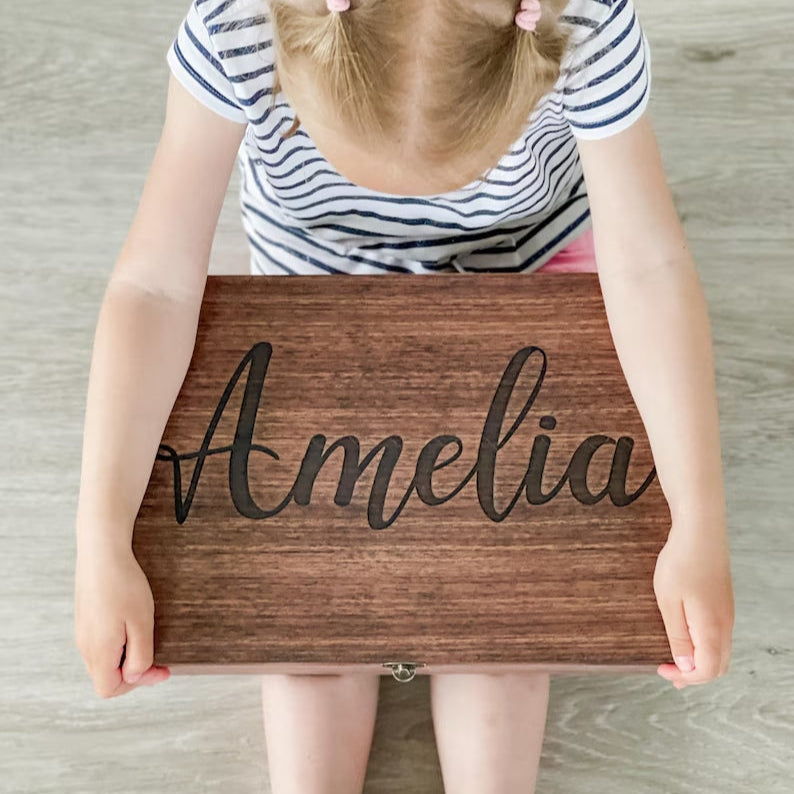 Child holding a wooden box with 'Amelia' engraved on it against a wooden floor background